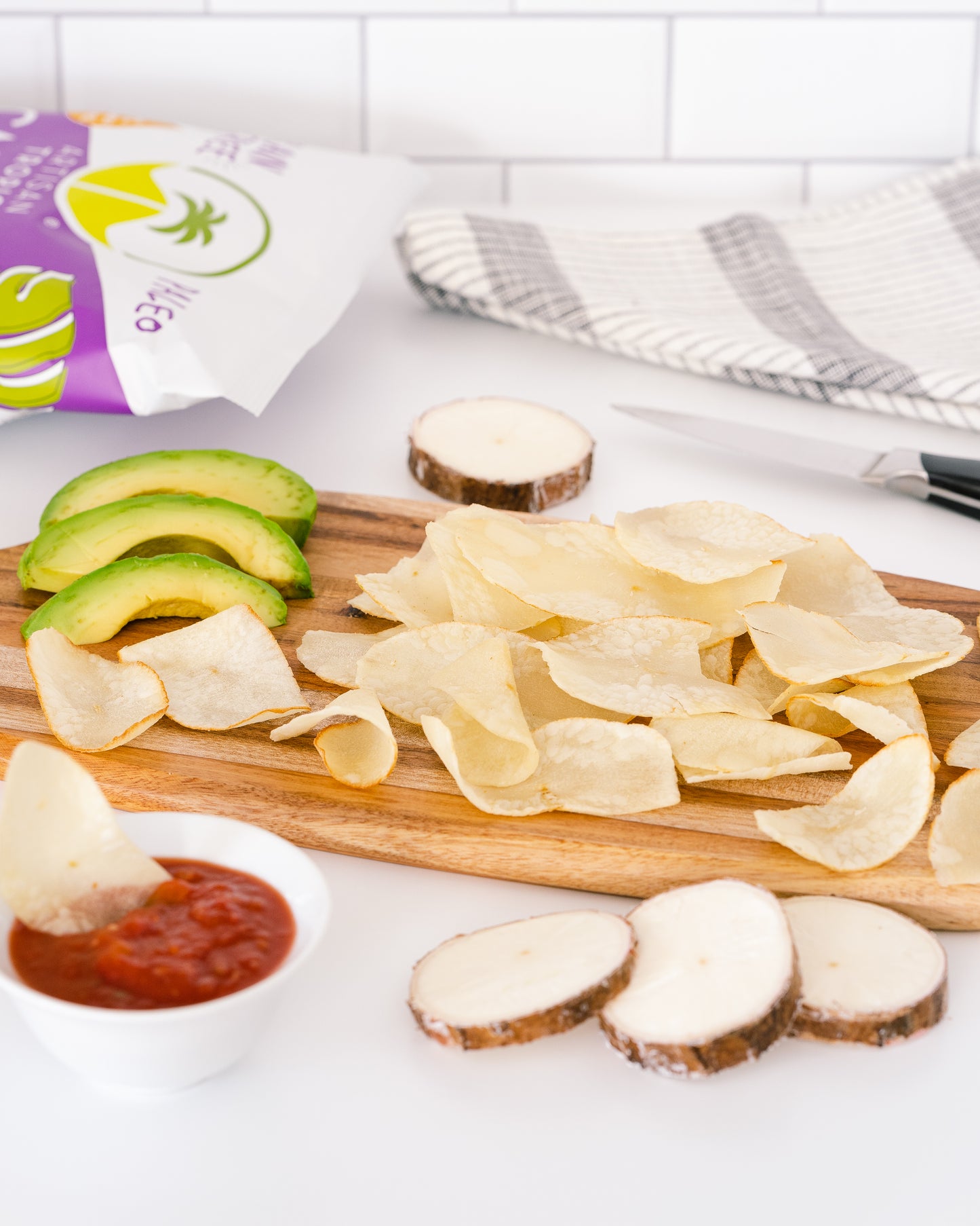 Wooden cutting board with sliced potatoes, avocado, and a small bowl of red sauce on a white surface.