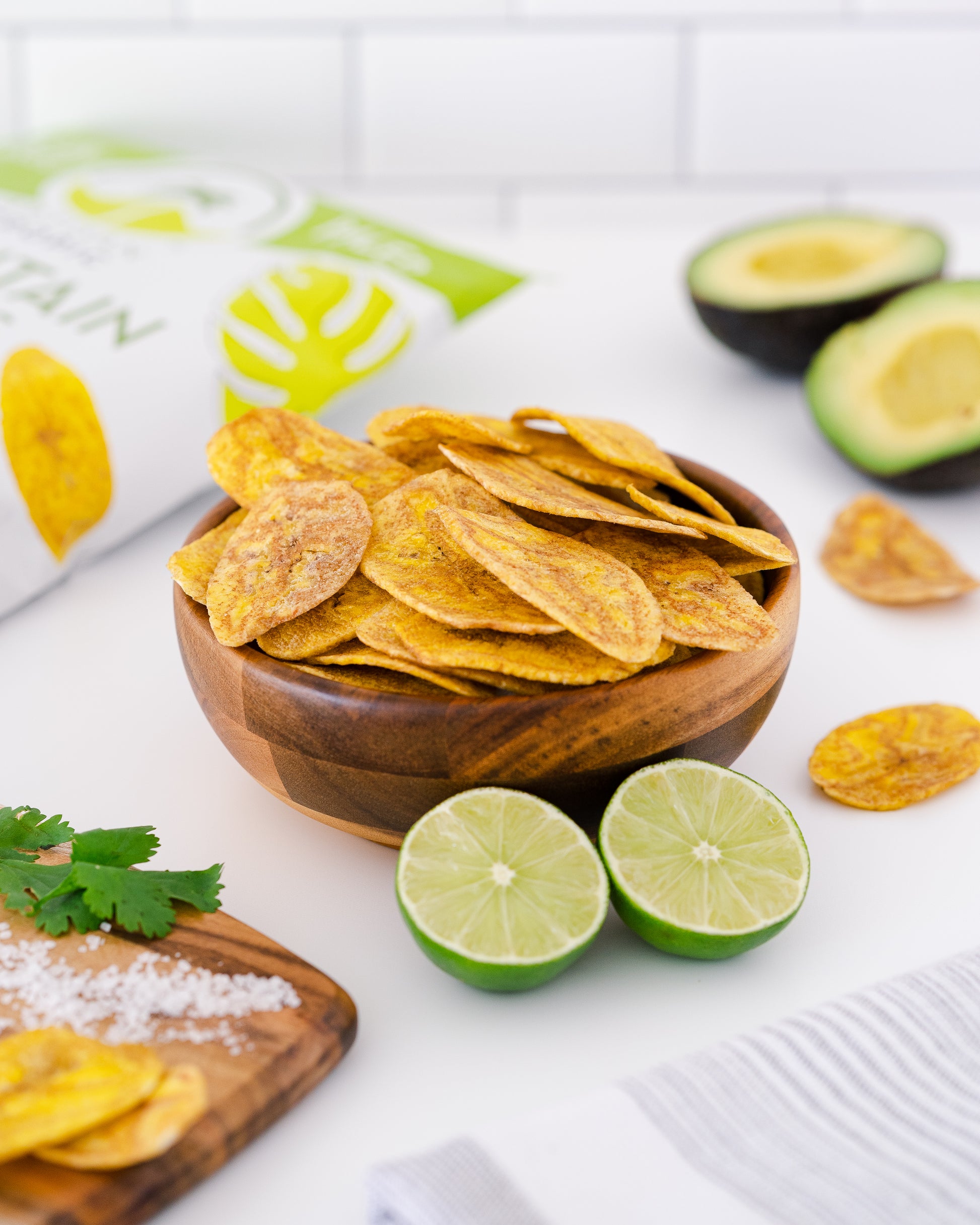 Wooden bowl of tortilla chips with limes and an avocado on a white surface